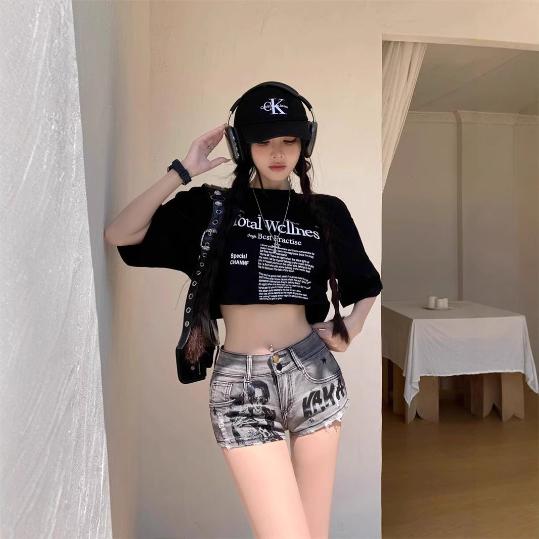 Person wearing a black crop top with text, denim shorts, and a black cap indoors.